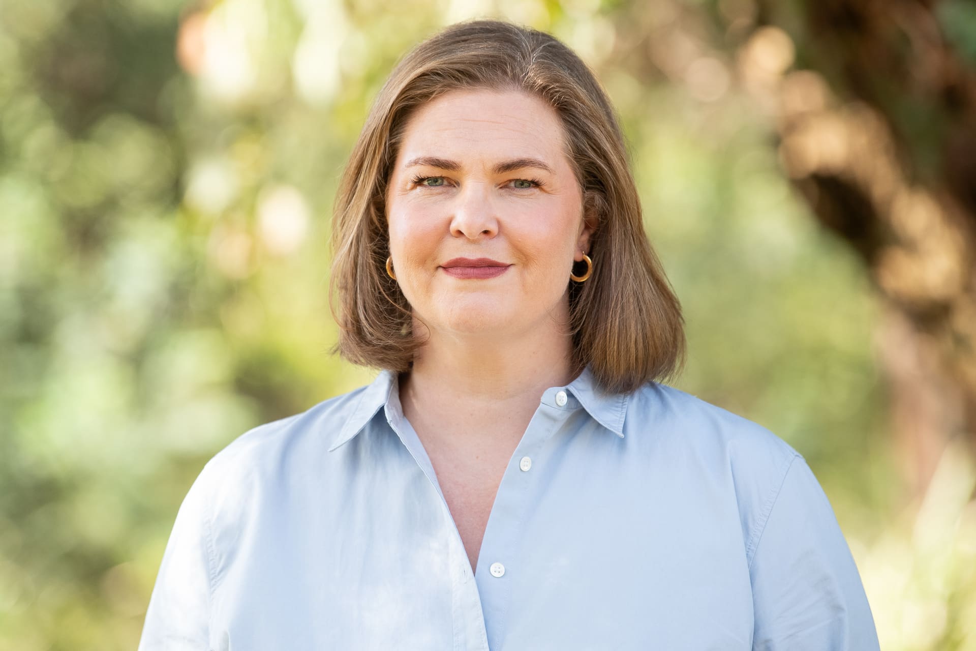 Outdoor headshot of woman in light blue linen shirt, with a soft smile to camera. The background is bright greenery.