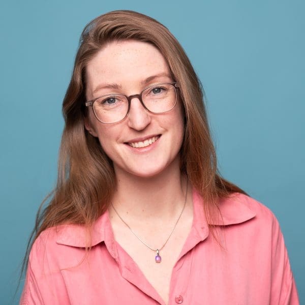 Headshot of Julia nance in a pink shirt on a blue background, smiling to camera.