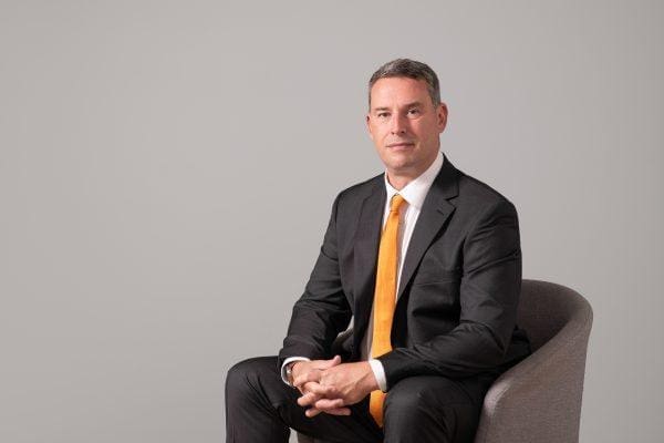 Formal corporate portrait of a man in suit and tie seated in an armchair on a light grey background.