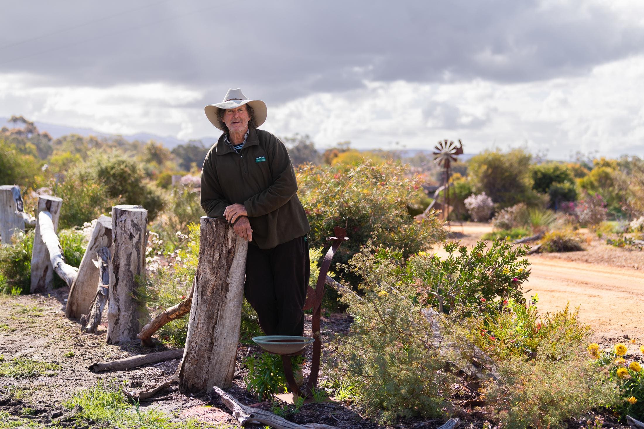 Phil Vaughan stands in his Pomonal garden, leaning on an old wooden fence post. He wears a wide brimmed hat and is surrounded by plants.