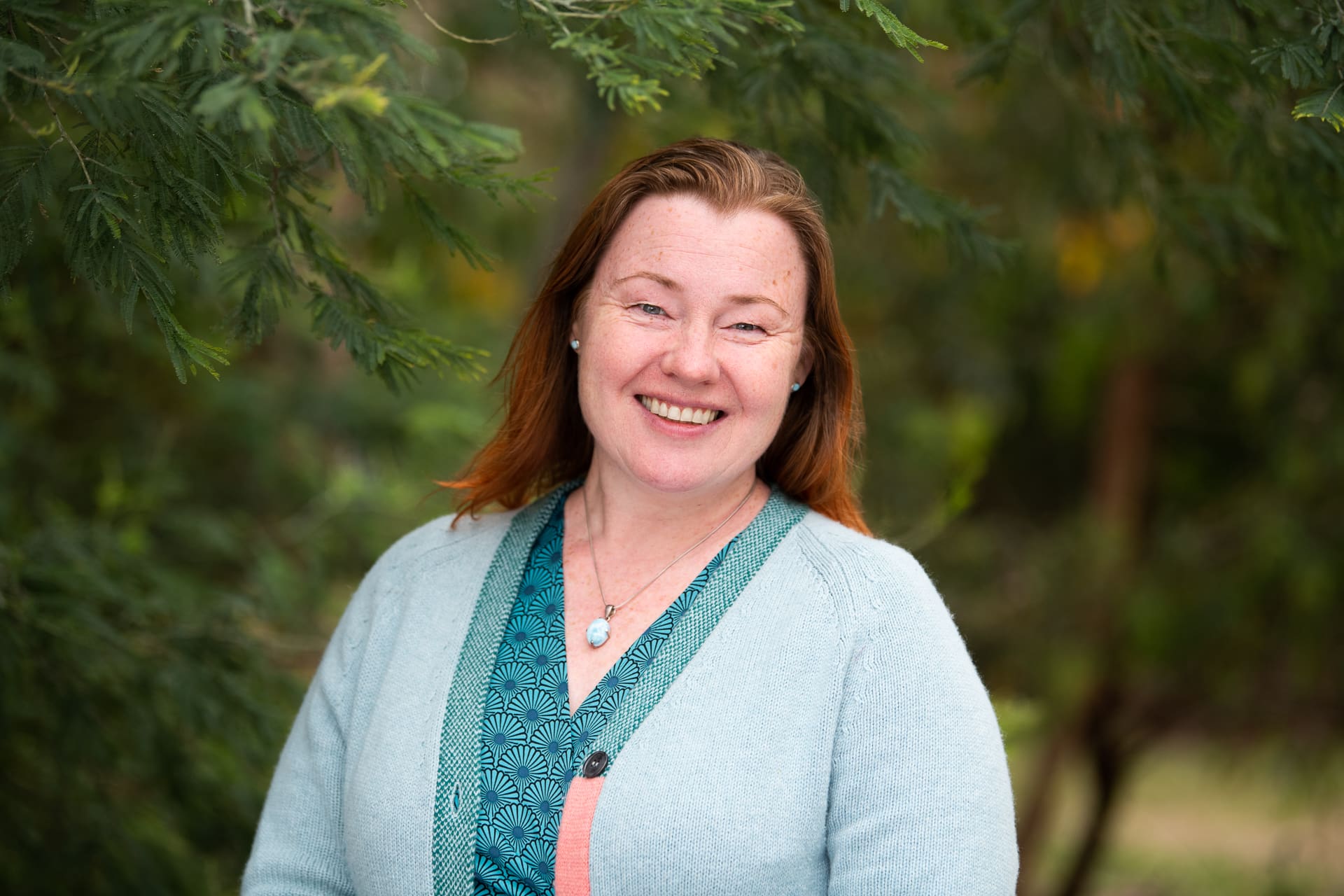 Outdoor headshot of woman smiling to camera with lush greenery background.