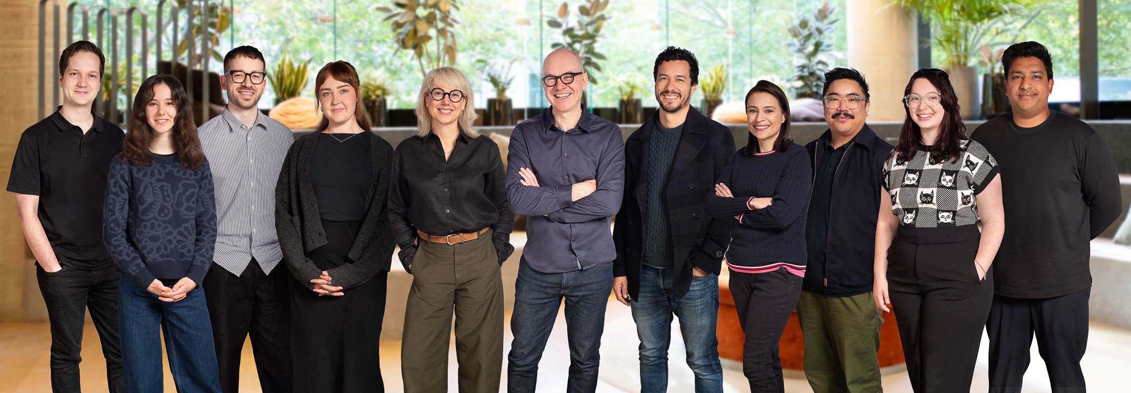 A team of 11 people stand in a staggered row, smiling to camera for a team portrait. They are in an large, warm office setting with plants behind them.