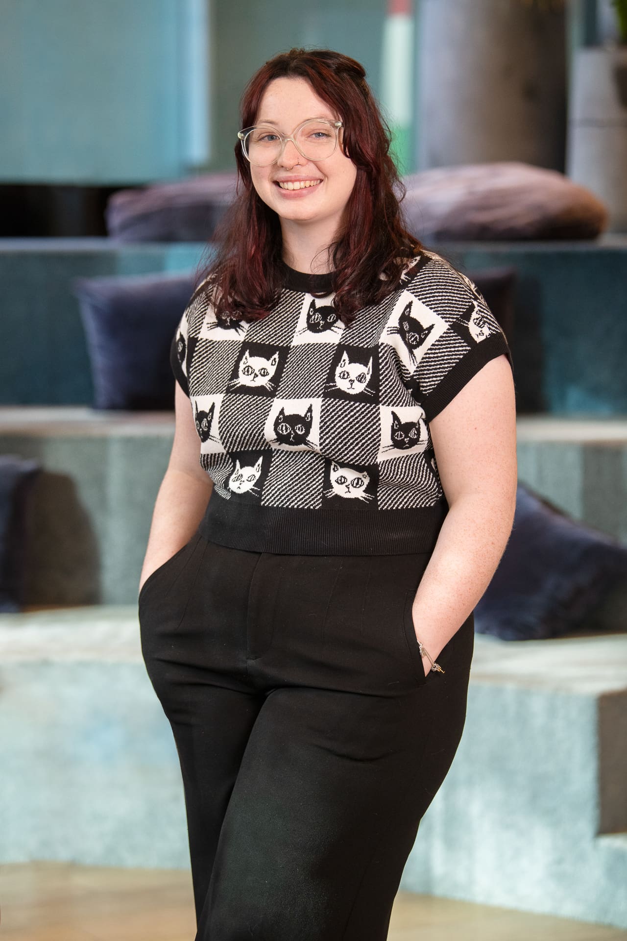 Woman stands in a coworking space setting, for soft portrait smiling to camera. She has her hands in her pockets and her top has black and white cats.
