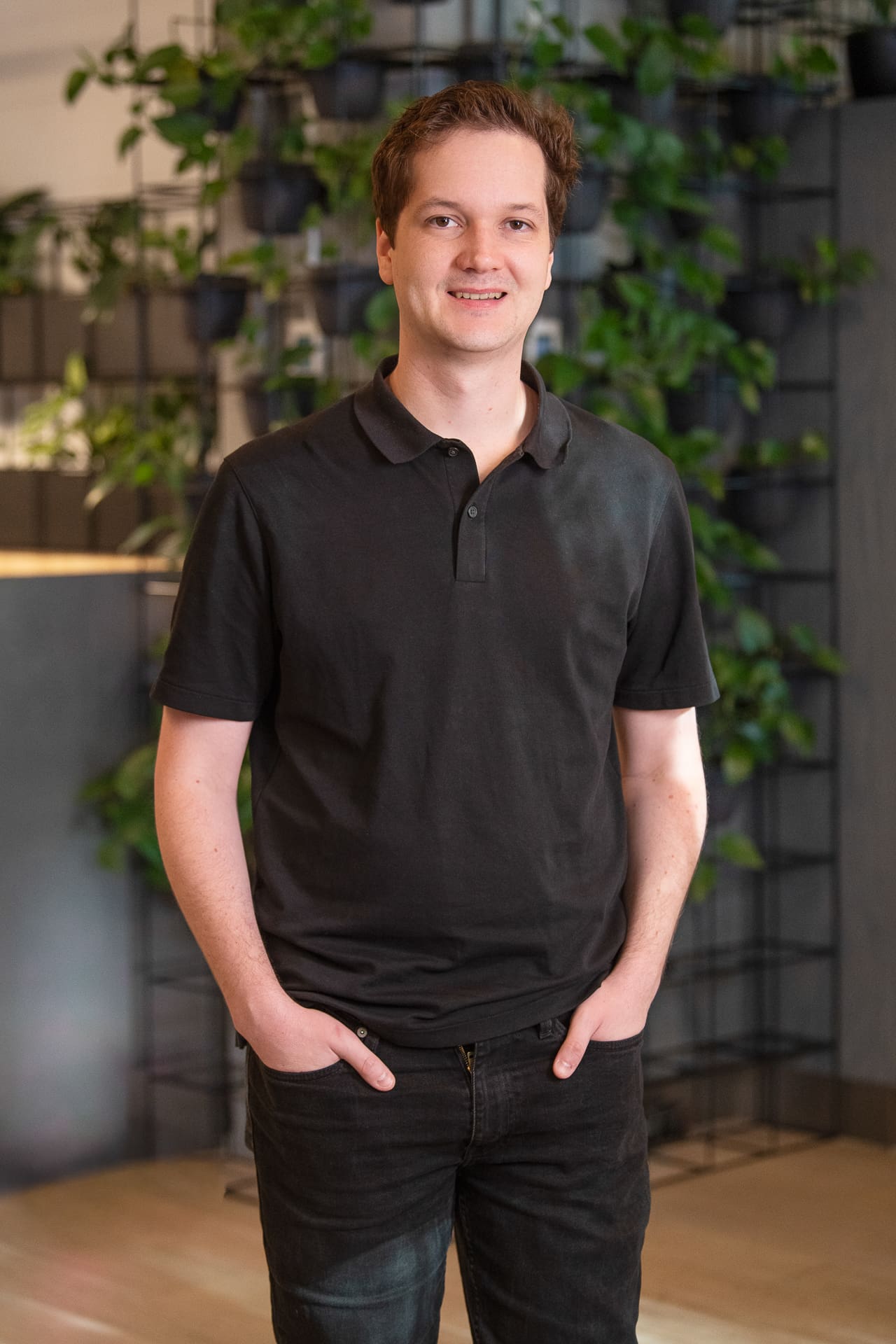 Man stands for relaxed company portrait in office setting, with plants behind him. He has his hands in his pockets.