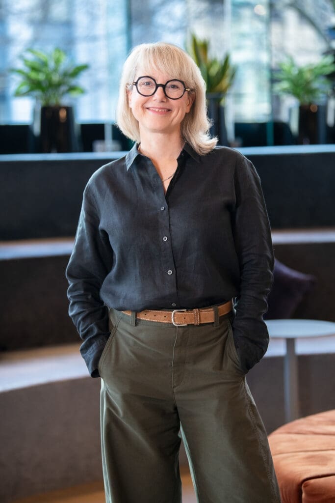 Woman stands in bright interior space smiling to camera, with relaxed pose.