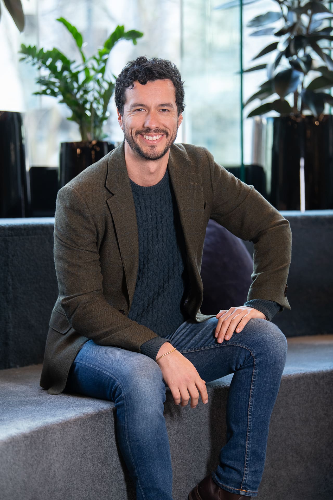 Man sits with plants behind him in large coworking space. He smiles to camera.