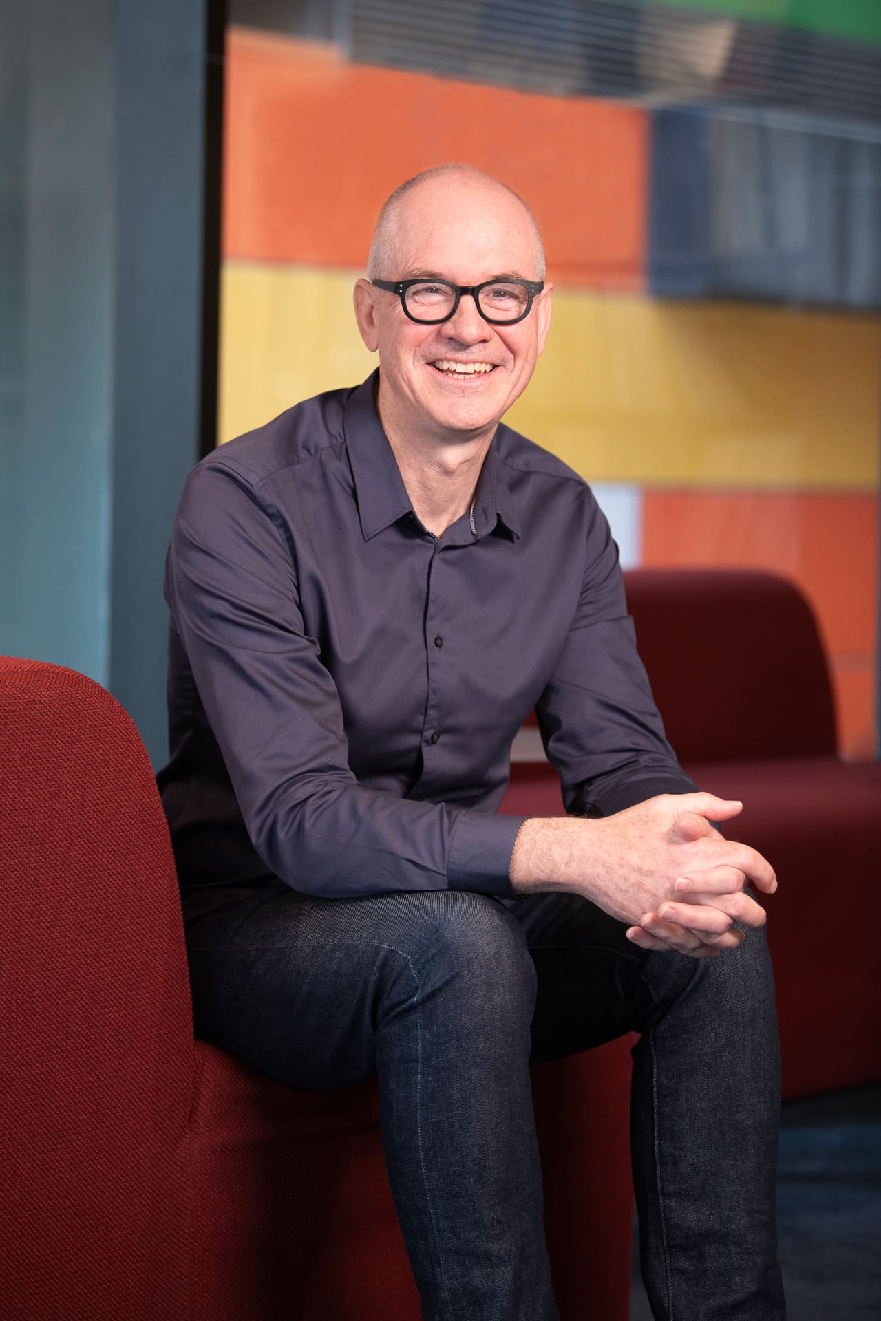 man smiles to camera in a seated pose, with colourful interior behind him for a relaxed company portrait.