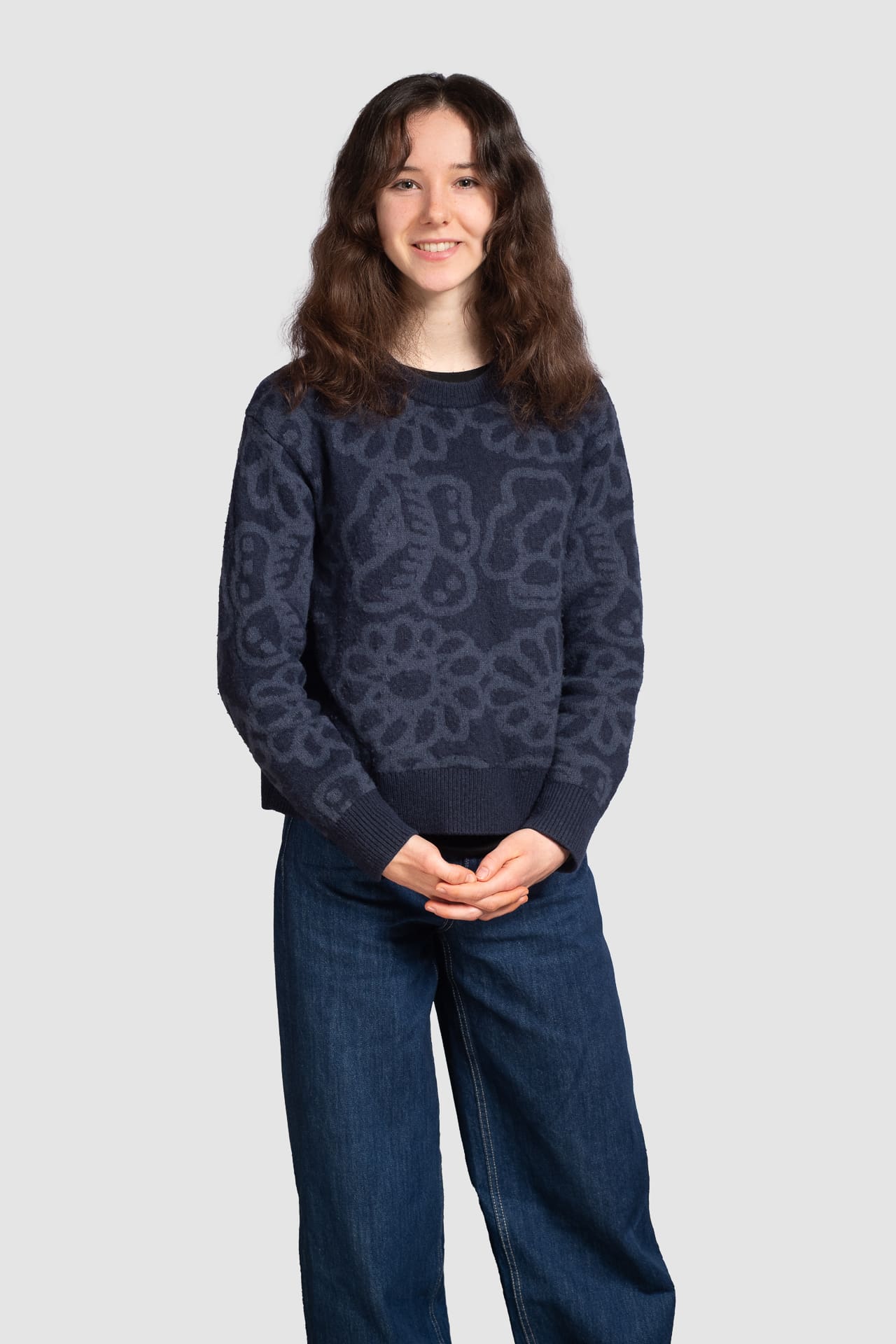 Woman stands on a light grey background for corporate portrait, smiling to camera with a relaxed pose.