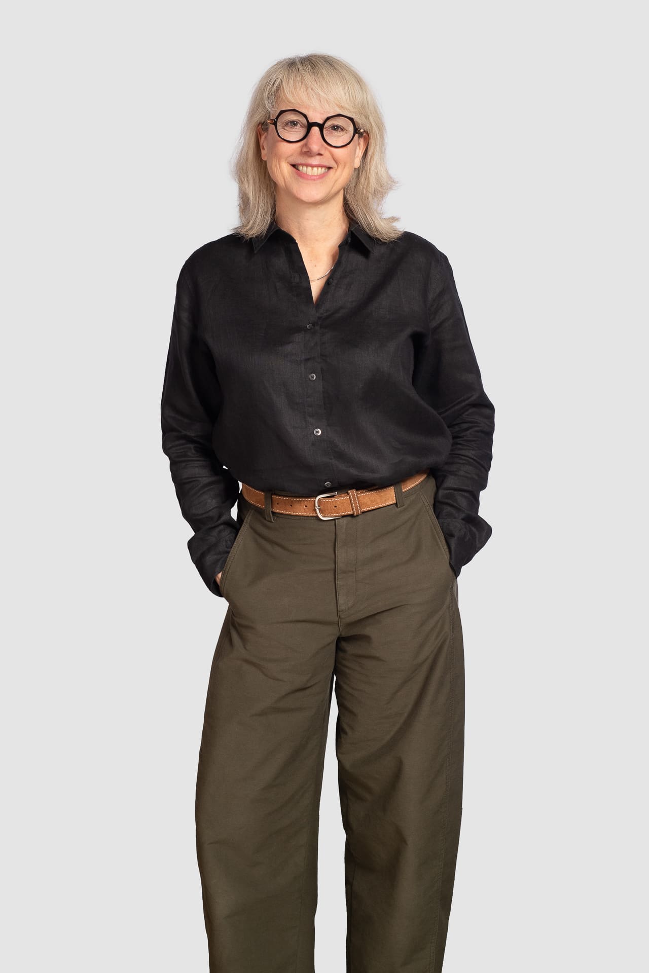 Woman stands on a light grey background, smiling and with hands in pockets for her corporate portrait.