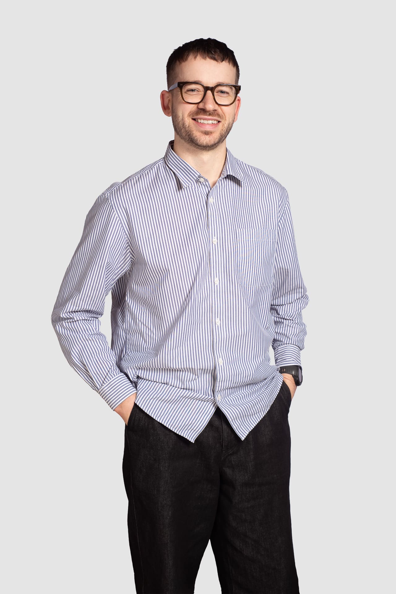 Corporate portrait on a light grey background of a man smiling to camera with his hands in his pockets.