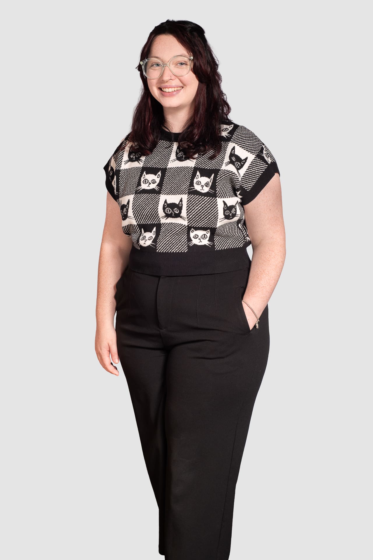 Woman stands for corporate portrait on light grey background. She smiles to camera with one hand in her pocket.