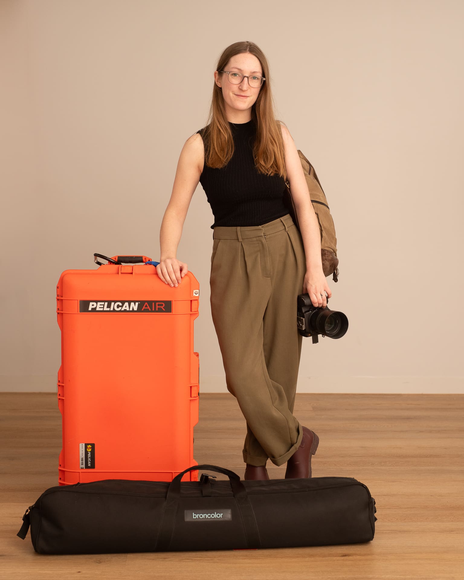 Julia stands looking to camera, holding her camera in one hand and rests her other hand on a suitcase holding her equipment. She wears a backpack and has a second light-stand bag at her feet.