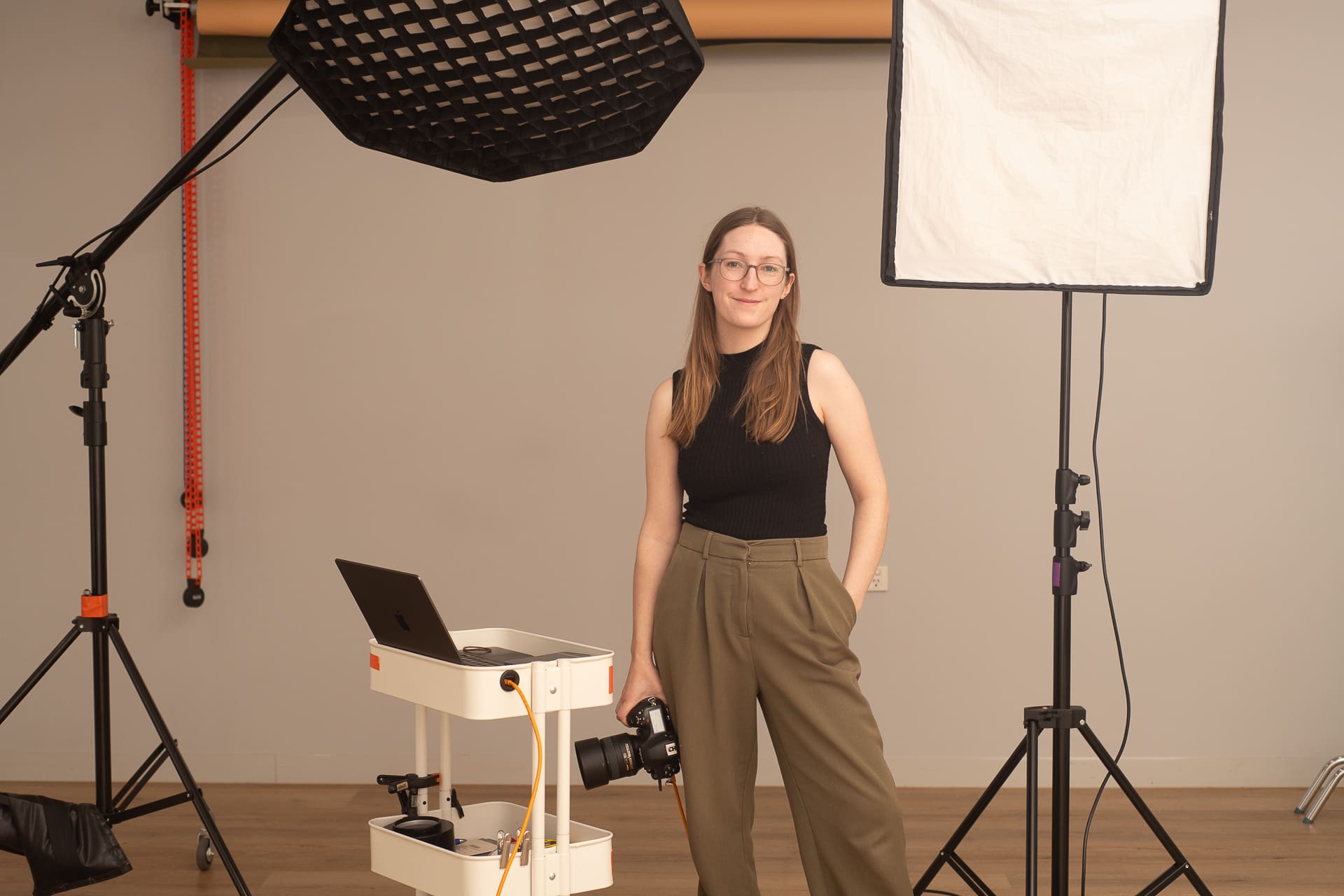 Julia stands looking to camera, holding her camera in one hand. There is lighting equipment around her and her laptop.